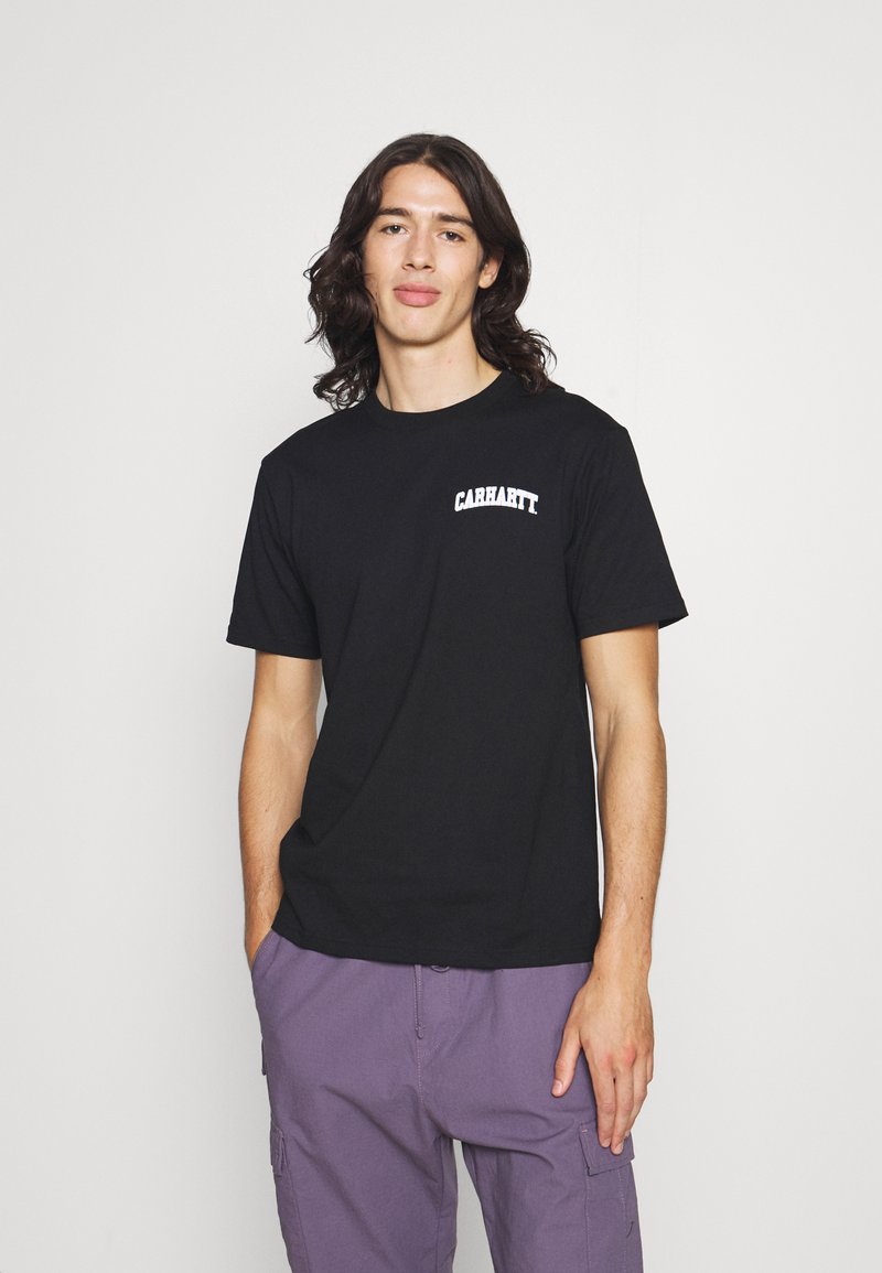 T-shirt en coton noir avec un logo "CARHARTT" blanc sur la poitrine gauche, col rond et manches courtes. Associé à un pantalon cargo violet.