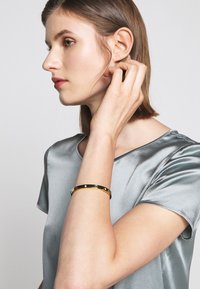 Bracelet doré avec des segments noirs alternés et des accents brillants sur une surface lisse. Porté au poignet, il complète un top en soie.