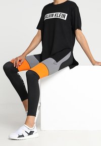Sort t-shirt med "CALVIN KLEIN" logo, parret med flerfarvede leggings med orange og grå paneler, og hvide sportssko.