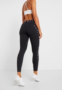 Black leggings with white branded text down the sides, made of stretchy material. Paired with a light gray sports bra and white sneakers.
