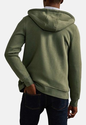 Homme portant un sweat à capuche vert olive, les mains près des poches, vu de dos sur un fond uni.