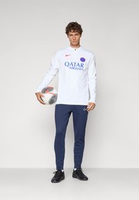 Bijela košulja dugih rukava s plavim logotipom "QATAR AIRWAYS", tamnoplave hlače i crne nogometne cipele, drži crno-bijelu nogometnu loptu.