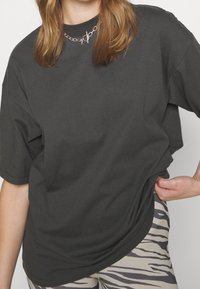 Persona con una camiseta negra holgada, collar de cadena de plata y leggings con estampado de cebra, con una mano tirando del dobladillo de la camiseta.