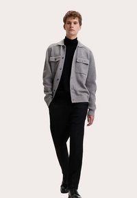 Grå stickad cardigan med två framfickor, stängs med knappar, bärs över en svart polotröja. Matchas med svarta byxor och skor.