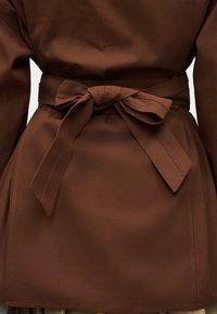 Σκούρο καφέ παλτό με φαρδιά υφασμάτινη ζώνη δεμένη σε φιόγκο στη μέση, που δείχνει λεπτομερείς ραφές και ομαλή υφή.