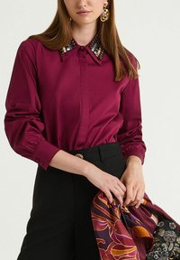Burgundy blouse with a structured collar featuring geometric sequin accents. Long sleeves with buttoned cuffs, paired with black trousers and a patterned scarf.