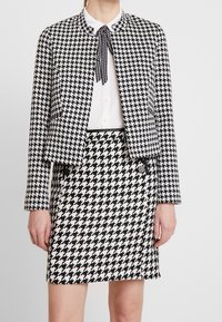 Blazer a motivo houndstooth bianco e nero con silhouette cropped, abbinato a una gonna houndstooth coordinata e una camicia bianca.