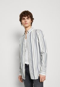 Gestreepte overhemden van lichte stof, met dunne blauwe en grijze strepen op een witte achtergrond. Gestyled met een effen witte onderhemd.