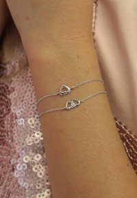Zilveren dubbele armband met twee hartvormige bedels en een kleine kraalaccent. De kettingen zijn fijn en delicaat met een gladde textuur.