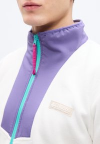 Μπουφάν από φλις με ψηλό γιακά, μισό φερμουάρ σε τιρκουάζ και ροζ, λευκό σώμα και μοβ λεπτομέρειες με λογότυπο στο στήθος.