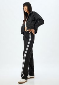 Schwarze cropped Jacke mit Reißverschluss, zwei Fronttaschen, über einem weißen Oberteil getragen. Schwarze Hose mit weißen Seitenstreifen und weißen Sneakers.