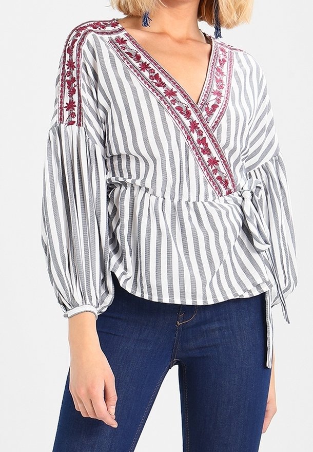 Femme portant un chemisier cache-cœur à rayures blanches et grises avec une broderie florale rouge sur les épaules et le décolleté, associé à un jean bleu foncé.