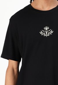 T-shirt in cotone nero con maniche corte, presenta un design floreale bianco nella parte sinistra del petto. Tessuto morbido, vestibilità rilassata.