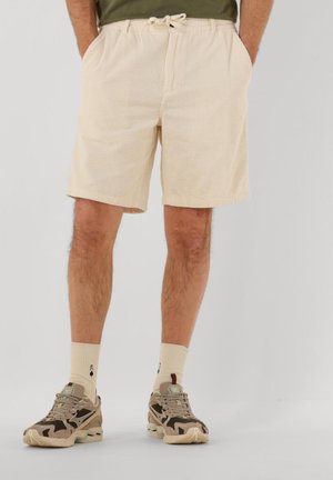 Pantalones cortos de pana beige con cintura ajustable con cordón, bolsillos laterales y un patrón acanalado texturizado. Modelo con zapatillas beige y marrones.