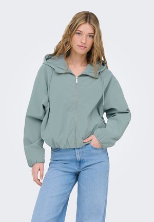 Mladá žena v svetlozelenom mikinovom zipsovom svetri s kapucňou a modrých rifliach stojaca s jednou rukou v vrecku proti jednofarebnému pozadiu.