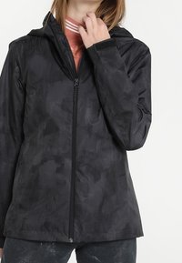 Person iført en sort lynlåjsjakke med subtilt camouflage-mønster og en pink bluse under, hånd nær kraven.