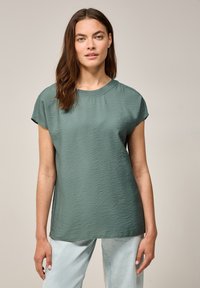 Top verde a maniche corte con scollatura rotonda, vestibilità rilassata e tessuto morbido e leggero. Indossato con jeans azzurri chiari.