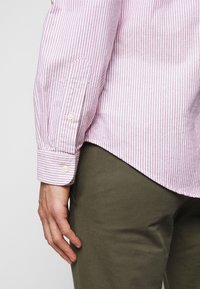 Skjorta med button-down krage i rosa och vita vertikala ränder, med manschetter som stängs med knappar och en slät tygkvalitet. Oljefärgad gröna byxor syns.