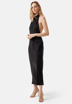 Black halter dress featuring an open back, knee-length cut, soft texture, and minimalist design. Paired with gold strappy heels.