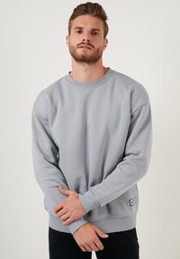 Sudadera gris de tela suave, con cuello redondo, corte holgado y puños acanalados. Pequeño parche de logo en la esquina inferior izquierda.