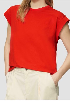 Mujer con una blusa sin mangas de color rojo brillante metida dentro de pantalones beige de cintura alta, con las manos en los bolsillos, vista desde el cuello hasta las caderas.