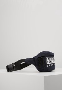 Borsa in tessuto blu scuro con cintura regolabile nera e logo bianco "Napapijri geographic" sul davanti, posizionata su una superficie bianca.