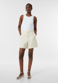 Weißes, geripptes Tanktop kombiniert mit beigen, strukturierten, hochgeschnittenen Shorts. Mit Seitentaschen, einem eleganten Design und minimalen weißen Riemchen-Highheels.