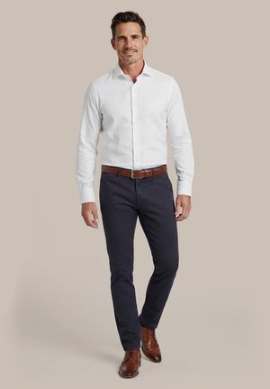 Mann geht vorwärts, trägt ein weißes Hemd, dunkelblaue Hose, braunen Ledergürtel und braune Anzugsschuhe vor einem beigen Hintergrund.