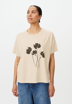 Kvinde iført en beige t-shirt med et sort blomsterdesign og blå jeans, stående mod en ensfarvet hvid baggrund.