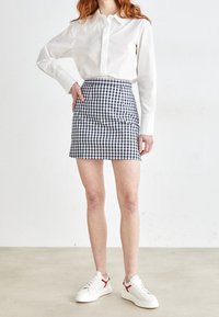 Weißes Button-Down-Hemd, kombiniert mit einem Minirock aus blau-weißem Gingham-Muster. Das Ensemble wird durch weiße Sneaker mit roten Akzenten ergänzt.