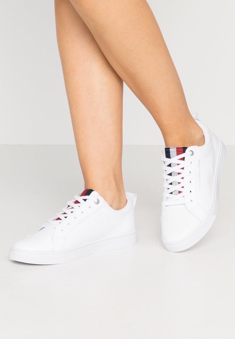 Baskets blanches avec une texture lisse, un design profil bas et un détail rayé contrastant sur la languette en rouge et bleu marine. Fermeture à lacets et semelle en caoutchouc.