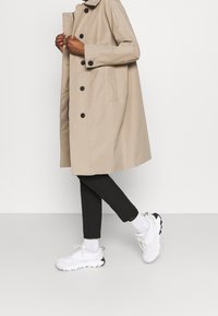 Persona con un abrigo trench beige, pantalones cortos negros, calcetines blancos y zapatillas gruesas blancas, de pie contra un fondo blanco liso.
