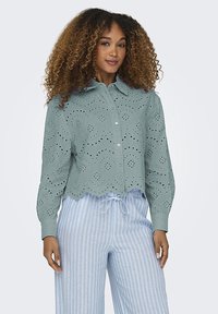 Groene blouse met oogjes, een golvende zoom en knoopsluiting, met ingewikkelde uitsnijdingen. Gecombineerd met lichtblauwe gestreepte broek.