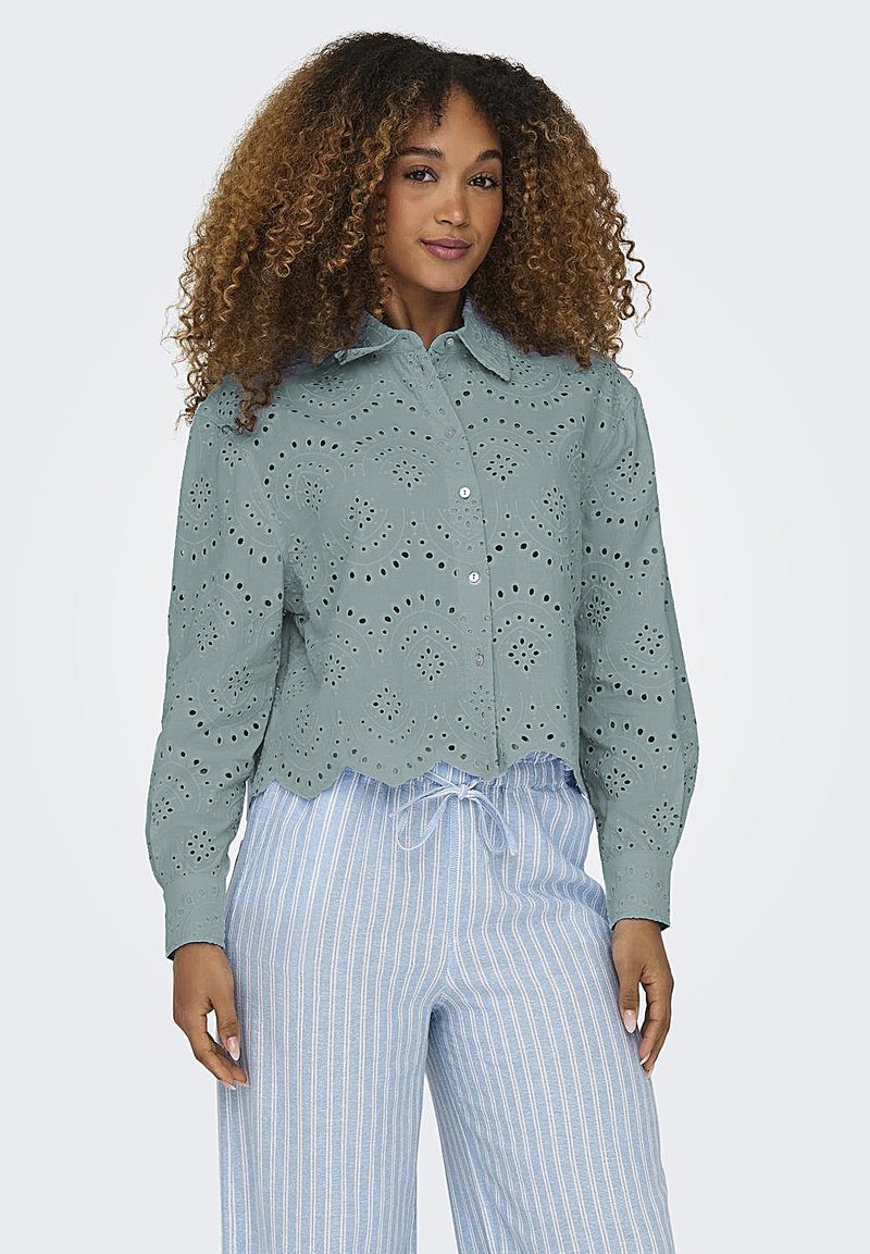 Groene blouse met oogjes, een golvende zoom en knoopsluiting, met ingewikkelde uitsnijdingen. Gecombineerd met lichtblauwe gestreepte broek.