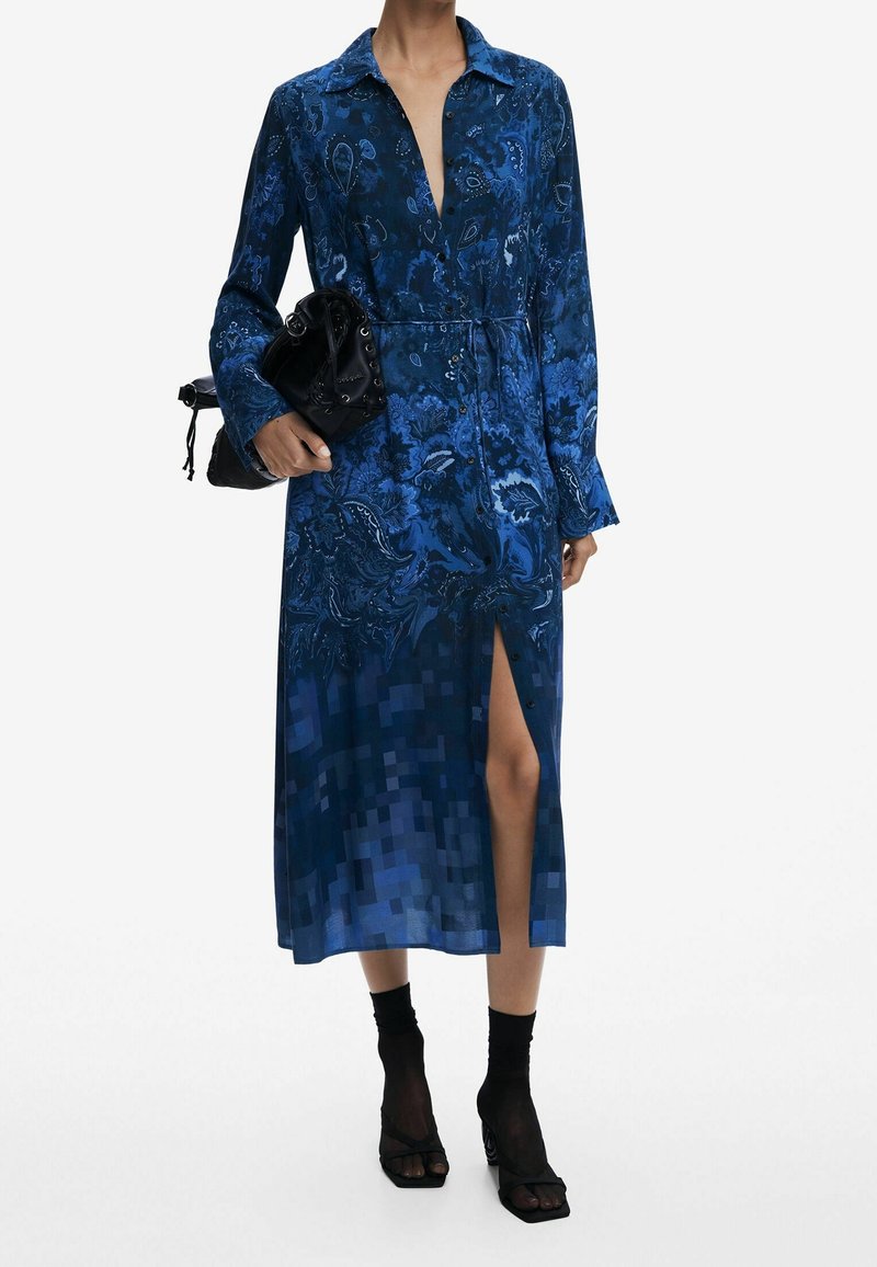 Femme portant une robe midi bleue à manches longues avec un motif et une fente à l'avant, tenant un sac à main noir et portant des sandales à talons noires avec des chaussettes.
