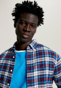 Camicia a quadri con controlli blu, rossi e bianchi sovrapposta a un maglione blu a tinta unita. Presenta colletto button-down e bottoni frontali.