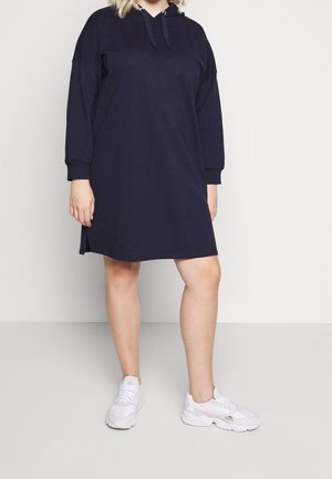 Person wearing a loose-fitting navy hoodie dress with long sleeves and white sneakers, standing against a plain light background.