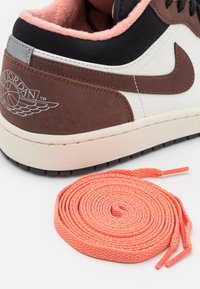 Baskets Air Jordan marron et blanches avec des accents noirs et des lacets couleur pêche enroulés sur une surface blanche.
