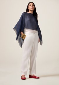 Poncho transparent bleu marine avec un col rond, associé à un pantalon large blanc et des ballerines rouges. Bracelet texturé en or au poignet.