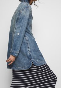 Veste en denim bleu clair avec une finition usée, dotée de manches longues, d'un col boutonné et d'un ourlet courbé. Portée par-dessus une jupe rayée.