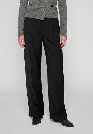 Personne portant un pantalon noir à jambes larges, un pull gris asymétrique boutonné et des chaussures noires à bouts pointus, debout devant un fond uni.