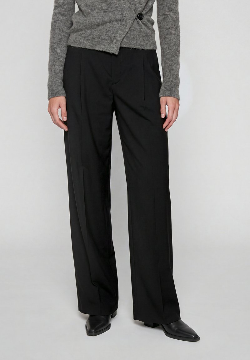 Personne portant un pantalon noir à jambes larges, un pull gris asymétrique boutonné et des chaussures noires à bouts pointus, debout devant un fond uni.