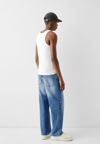 Canotta bianca senza maniche abbinata a jeans a gamba larga blu. Il modello indossa un cappellino nero e sneakers bianche, con dettagli di cucitura e due tasche posteriori.