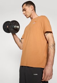 Brun bomull t-skjorte med korte ermer og rund hals, med en liten rød Puma-logo på brystet, kombinert med svarte shortser.
