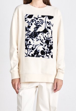 Woman wearing cream long-sleeve sweatshirt with black and white floral square design on front and matching light-colored pants.