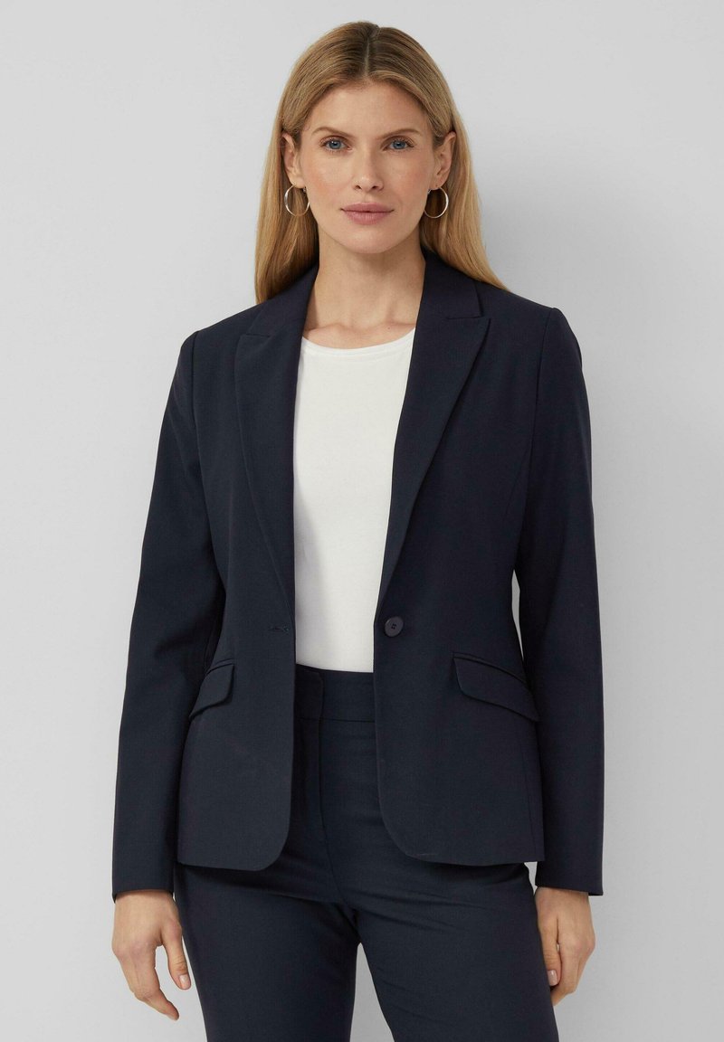 Jūrinės spalvos švarkas su pritaikyta iškirpte, su vieno mygtuko užsegimu, dviem priekinėmis kišenėmis ir nešiojamomis atvartomis. Dėvimas ant balto viršutinio rūbo.