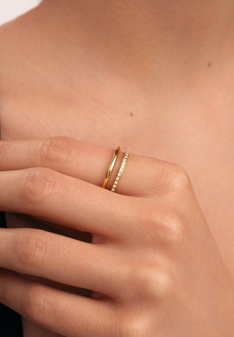Dos anillos en una mano: un aro de oro liso y un aro de oro engastado con pequeñas piedras transparentes. La mano tiene un tono de piel natural y texturas visibles.