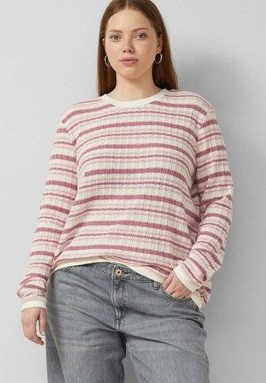 Jeune femme aux cheveux raides portant des boucles d'oreilles créoles, un pull rayé blanc et mauve, et un jean gris avec une main dans la poche.