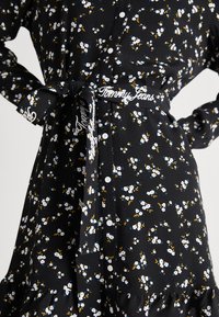 Abito nero floreale con motivi di fiori bianchi e blu, apertura con bottoni frontale, maniche lunghe e cintura in tessuto con la scritta "Tommy Jeans" in bianco.
