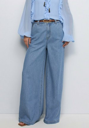 Blouse bleu clair à volants rentrée dans un jean bleu taille haute à jambes larges, avec une ceinture marron et des sandales ouvertes.